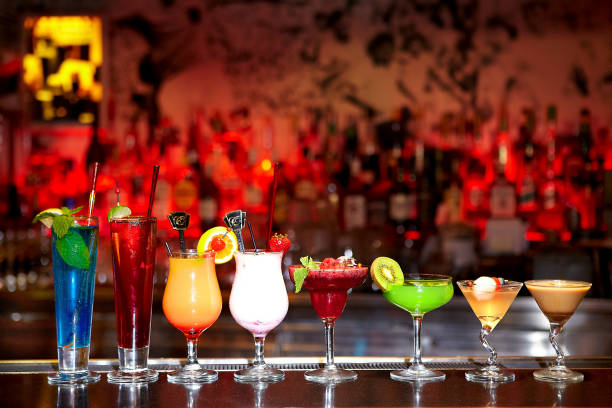 Assortment of eight homemade cocktails placed horizontally on a counter in a bar with a blurred background
